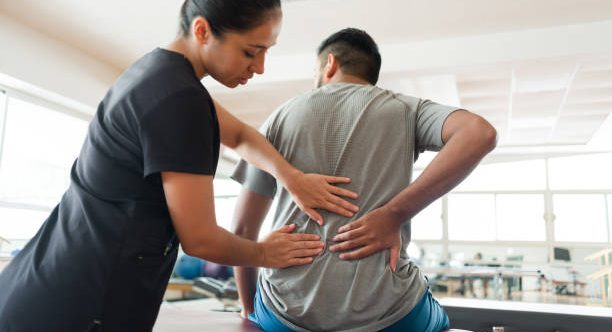 A female massage therapist standing behind a male patient and massaging his back with both hands.